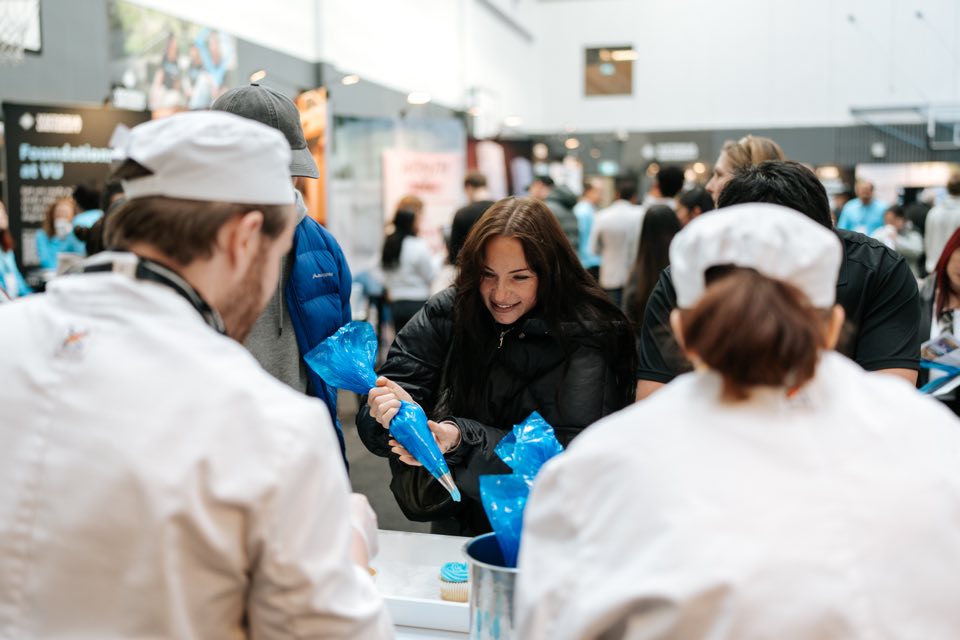 A visitor pipes blue icing onto a cupcake alongside VU cookery students in chef whites.