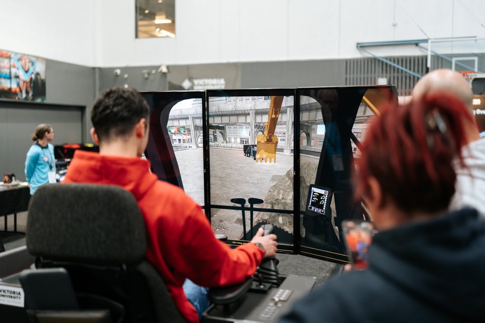 A visitor works the controls of VU’s excavator simulator while others watch the screens.