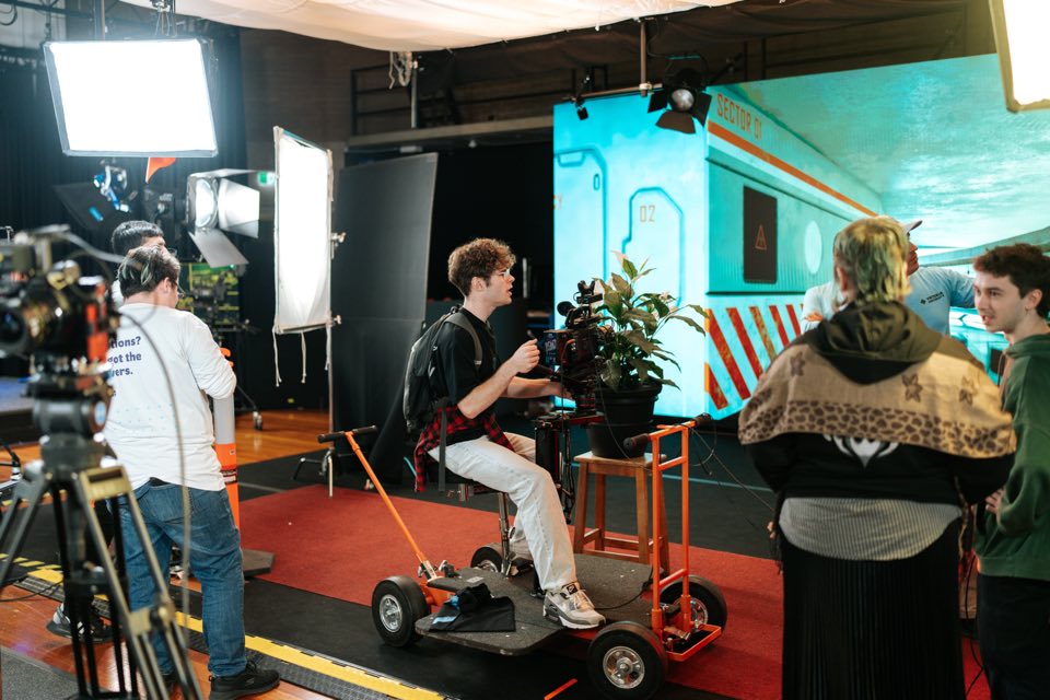A student operates a camera on a dolly in VU’s film studio in front of a lit set.